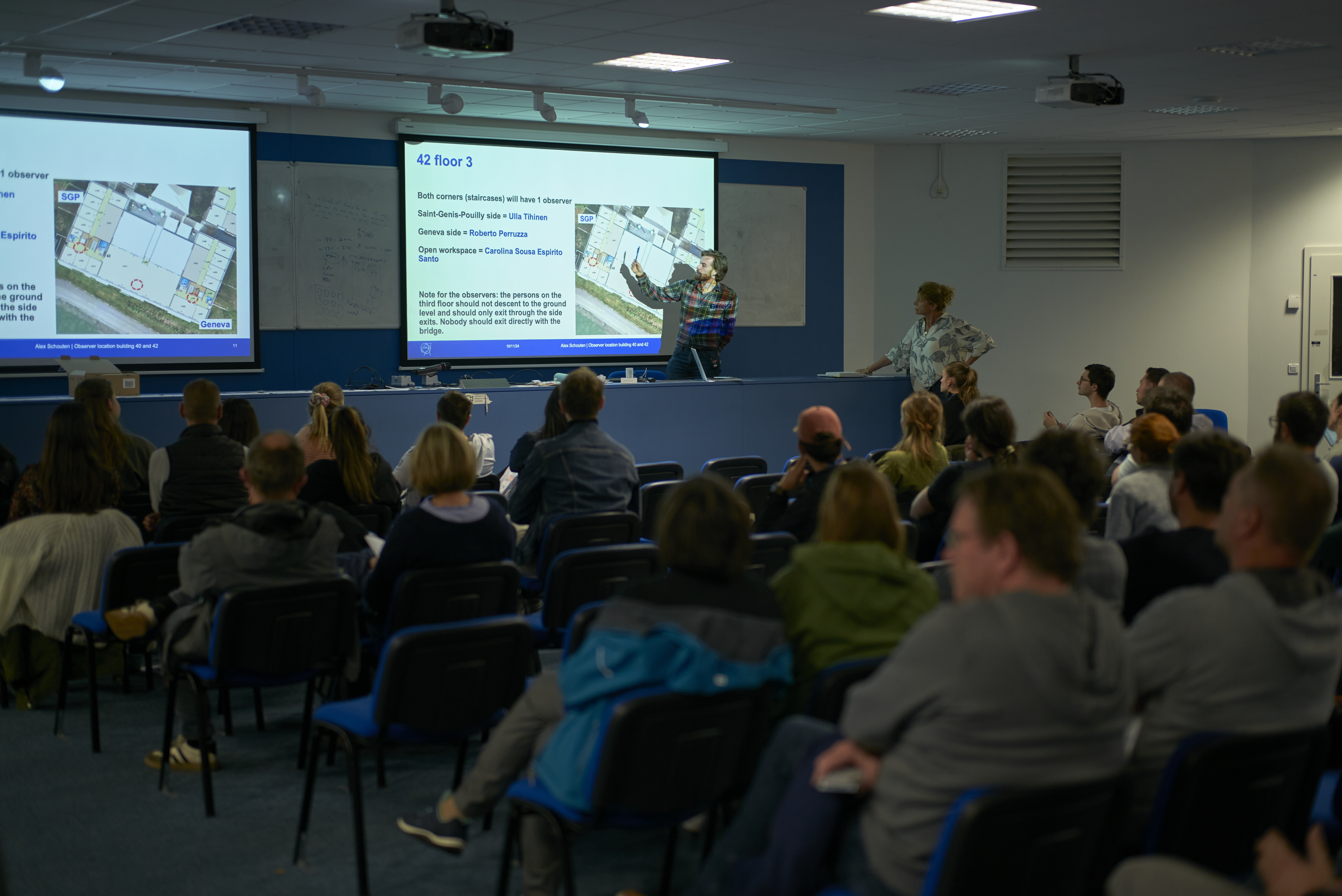 A large auditorium full of volunteers listens to the organisers, with a detailed map and schedule on the board.